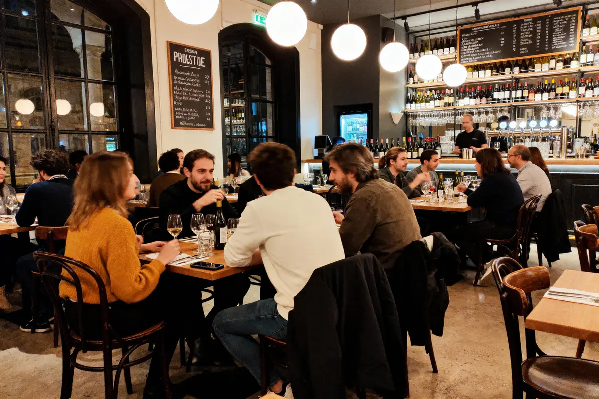 Des personnes dînent dans un restaurant chaleureux avec des tables en bois et un bar en arrière-plan.