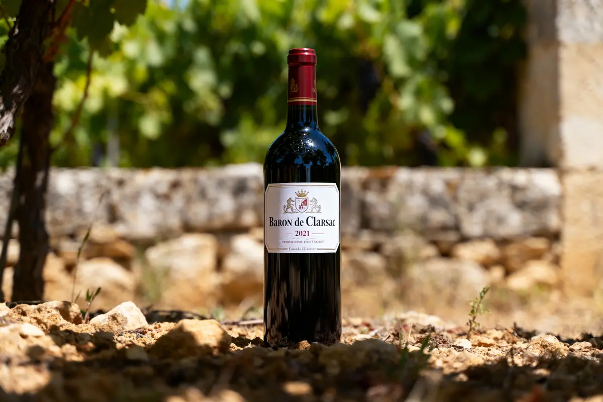 Bouteille de vin rouge Baron de Clarsac 2021 posée sur terre, avec vignoble en arrière-plan.