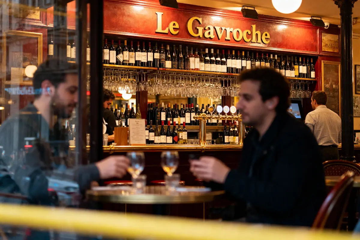 Deux personnes discutent dans un bar appelé "Le Gavroche", ambiance chaleureuse avec bouteilles et verres.