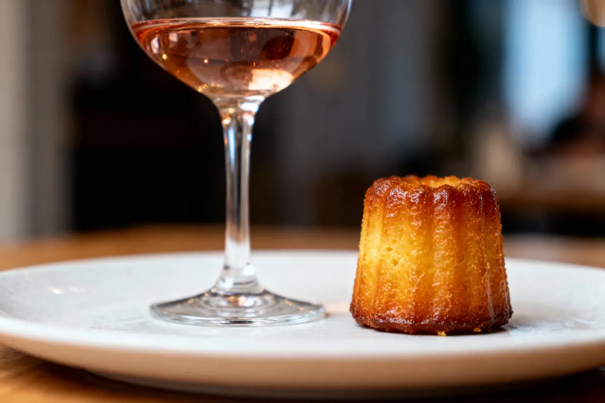 Verre de vin rosé et cannelé doré sur une assiette blanche, posé sur une table en bois.