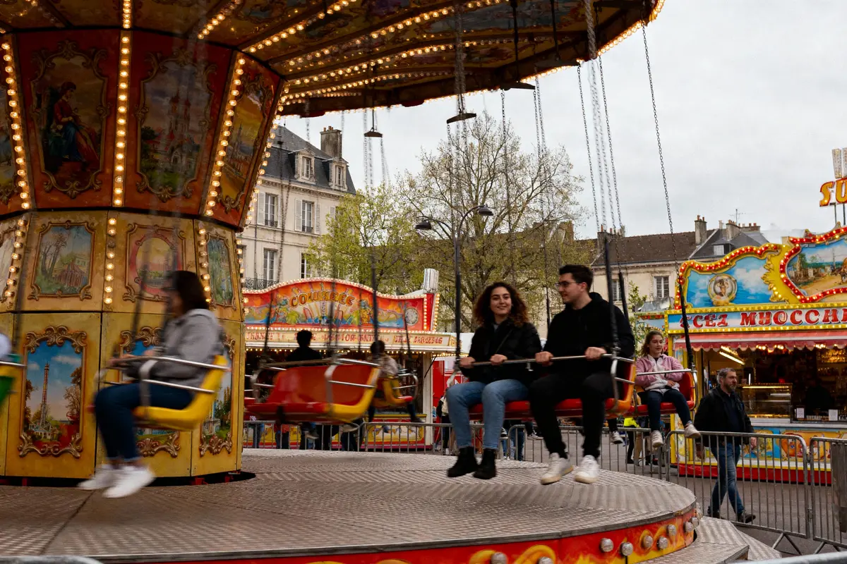 Manège avec personnes assises, bâtiments en arrière-plan, ambiance de fête foraine colorée et animée.