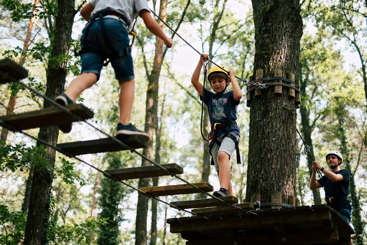 Deux enfants avec casques traversent un parcours d'accrobranche en forêt, encadré par un instructeur.